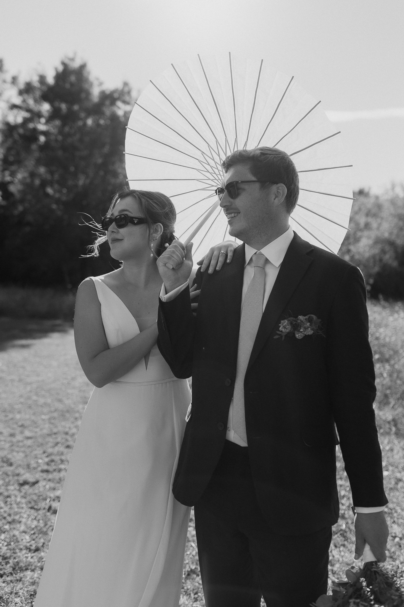 Black and white photo of a bride and groom wearing sunglasses in France on a field holding on to each other carrying a paper umbrella to shade from the sun