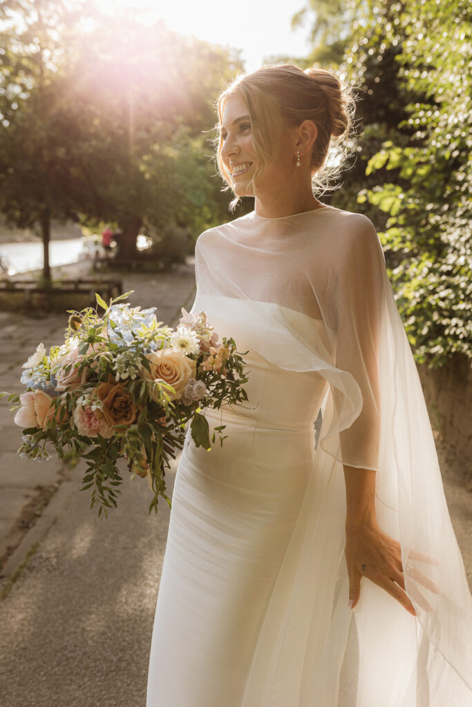 Colour image of a bride at sunset by York River it is golden hour and she is wearing a floaty cape and carrying a bouquet